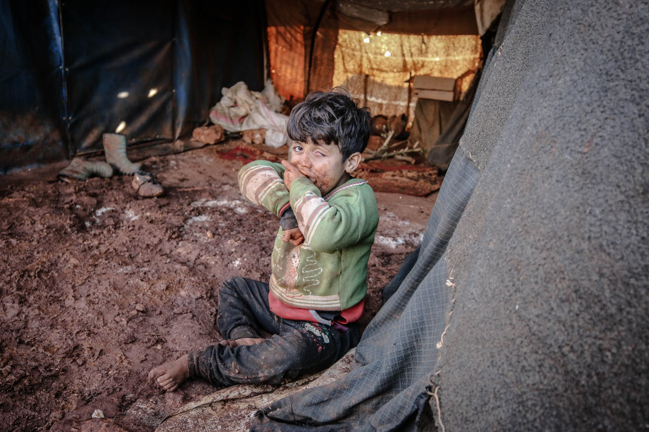 A young child with a dirty face sits in a refugee camp tent in Idlib, Syria.